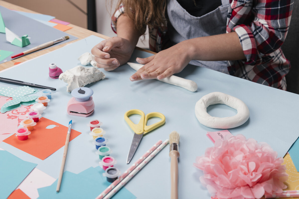 A person shaping air-dry clay on a craft table surrounded by paint and supplies