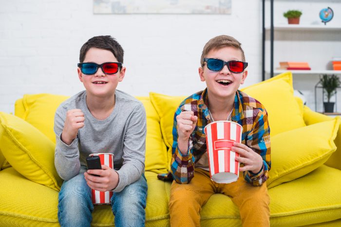 Two boys wearing 3D glasses on a yellow sofa holding tubs of popcorn