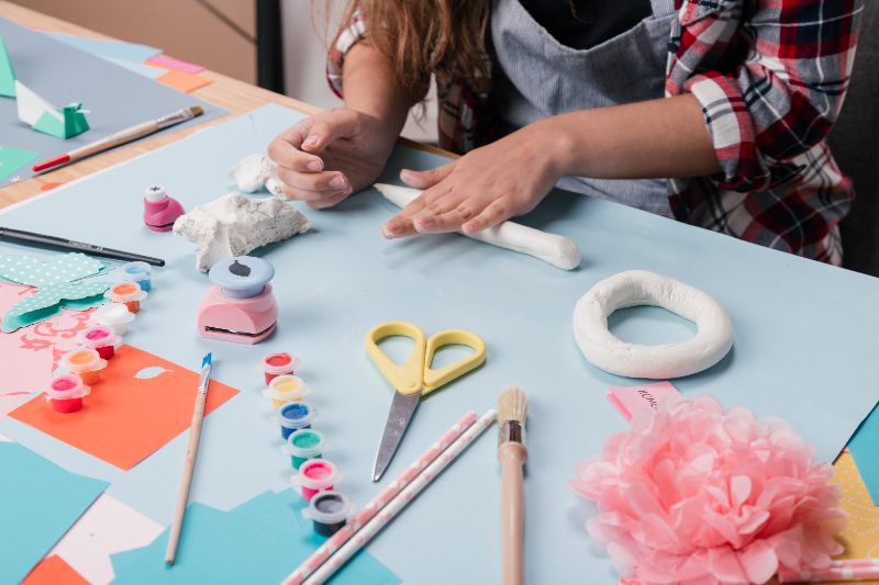 Hands shaping clay at a craft table covered in paints, paper and brushes